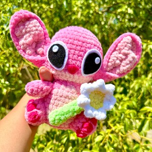 May include: A pink crocheted plush toy resembling a cartoon alien holding a white crocheted flower. The toy has large black eyes with white outlines and a pink nose.