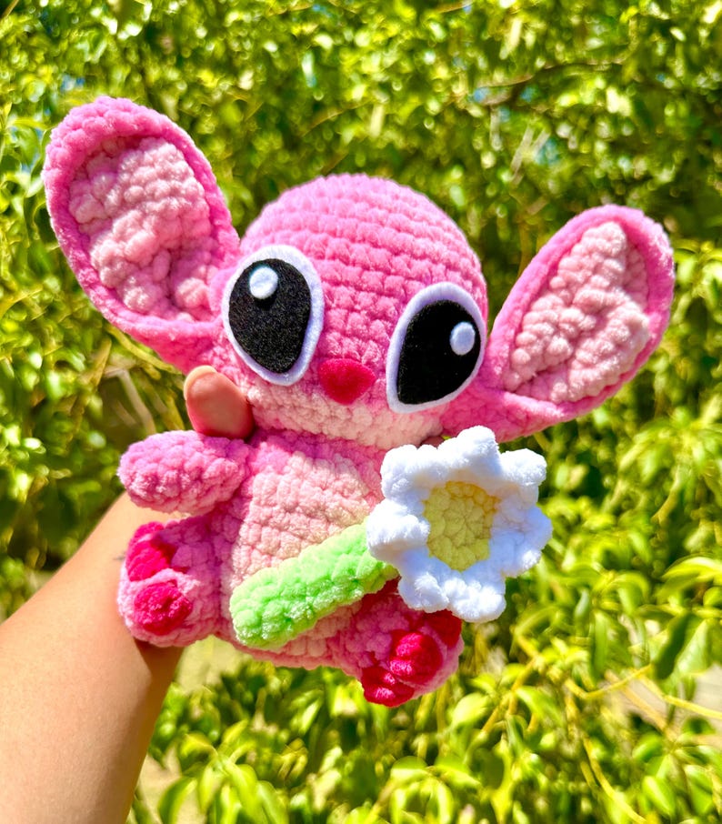 May include: A pink crocheted plush toy resembling a cartoon alien holding a white crocheted flower. The toy has large black eyes with white outlines and a pink nose.