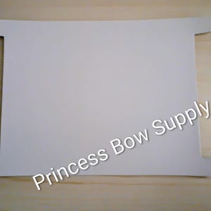 May include: A white, rectangular craft supply with four small tabs extending from the corners. The text "Princess Bow Supply" is printed diagonally across the surface. The item is set against a light wood grain background.