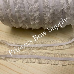 May include: A roll of ruffled elastic ribbon in a light beige color. The ribbon is partially unrolled, revealing its delicate, textured edge. The text "Princess Bow Supply" is printed on the image. This ribbon is suitable for various crafting projects.