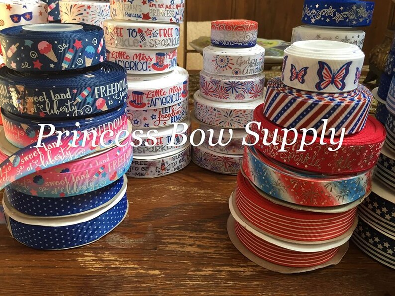 Pu&ograve; includere: Una collezione di nastri a tema patriottico in vari colori, tra cui rosso, bianco e blu. I nastri presentano disegni come stelle, strisce, fuochi d'artificio e testi come "Freedom" e "Little Miss America". Il testo "Princess Bow Supply" &egrave; anche visibile.
