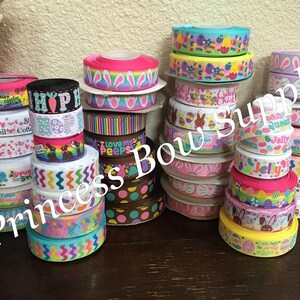 Pu&ograve; includere: Una collezione di nastri colorati a tema pasquale. I nastri presentano vari motivi, tra cui uova di Pasqua, coniglietti e testi come "Hoppy Easter" e "Jelly". I nastri sono impilati in rotoli, mostrando una variet&agrave; di colori e design.