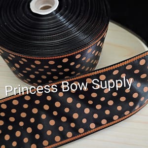 May include: A spool of black satin ribbon featuring a repeating pattern of tan polka dots and thin orange stitched edges. The text Princess Bow Supply is printed in white across the center of the ribbon, which is wound into a circular roll.