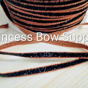 May include: A spool of narrow ribbon featuring a central black glittery stripe bordered by thin copper-colored edges. The text Princess Bow Supply is overlaid across the center of the image. The ribbon is wound into a tight circular coil.