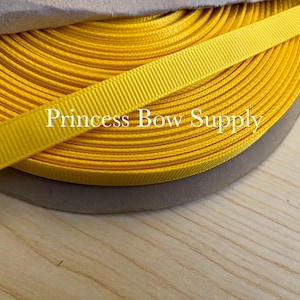 May include: A spool of bright yellow grosgrain ribbon with a ribbed texture. The ribbon is partially unrolled, revealing the coiled form. The text "Princess Bow Supply" is visible on the ribbon. The ribbon is likely used for crafting and embellishments.