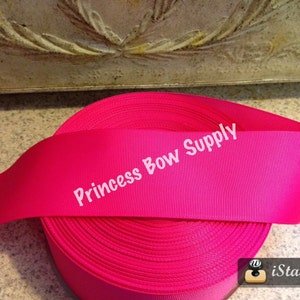 May include: A roll of bright pink grosgrain ribbon. The ribbon is labeled "Princess Bow Supply" in white text. The ribbon is tightly wound, showing the texture of the fabric. The background is a neutral color.