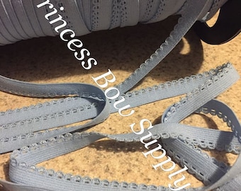 Può includere: Un rotolo di nastro elastico azzurro con un bordo smerlato. Il nastro è srotolato a sezioni, mostrando la trama e i dettagli. Il testo "Princess Bow Supply" è stampato sul rotolo.