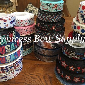 Pu&ograve; includere: Una collezione di rocchetti di nastro decorativo con vari disegni patriottici. I nastri presentano motivi con stelle, strisce, occhiali da sole e le scritte "USA" e "Freedom". L'immagine include il testo "Princess Bow Supply".