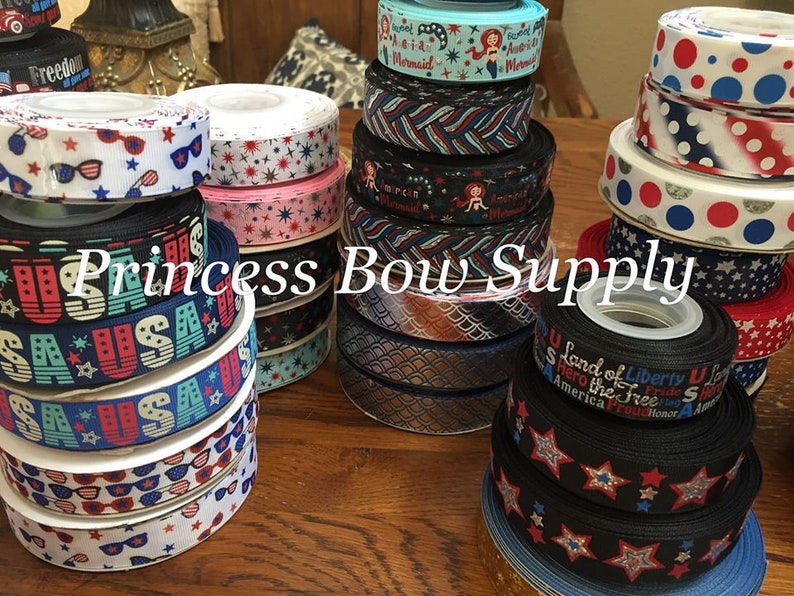 Pu&ograve; includere: Una collezione di rocchetti di nastro decorativo con vari disegni patriottici. I nastri presentano motivi con stelle, strisce, occhiali da sole e le scritte "USA" e "Freedom". L'immagine include il testo "Princess Bow Supply".