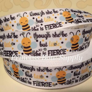 May include: White ribbon with a repeating pattern of smiling cartoon bees, blue and orange accents, and the text "though she be but little she is FIERCE". The ribbon has a glittery finish.