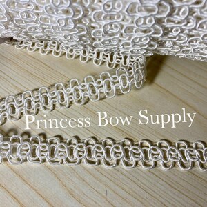 May include: A roll of white decorative trim with a looped design. The trim is made of a soft, woven material and is displayed on a light wood surface. The text "Princess Bow Supply" is visible on the surface.