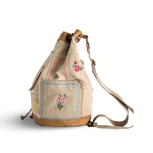 May include: A beige canvas backpack with a drawstring closure and leather accents. The backpack is decorated with embroidered floral patches in pink, yellow, and blue.