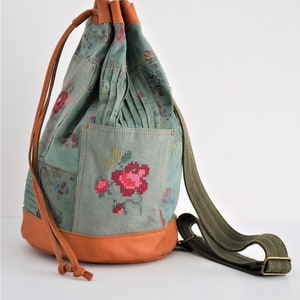 Op de afbeelding: Een groene canvas trekkoordtas met een bloemenpatroon en geborduurde rozen. De tas heeft een bruine leren afwerking en een groene canvas schouderband.