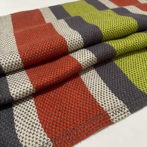 May include: A close-up shot of a folded textile with a woven design. The fabric displays stripes in rust orange, grey, off-white, and lime green. The texture of the material is visible, indicating a robust construction.