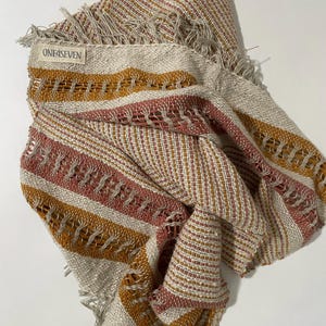 May include: A woven textile in beige, rust, and gold stripes, with a fringed edge. The label reads "ONE4SEVEN". The design incorporates open weave sections and solid stripes, creating a textured appearance. The textile is suitable for a scarf or shawl.