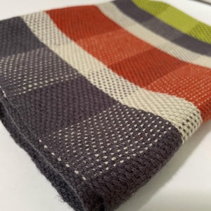May include: Close-up of a folded, woven textile with a checkered pattern. The design features stripes of dark gray, off-white, orange, and green. The fabric has a textured appearance, with small, evenly spaced holes.