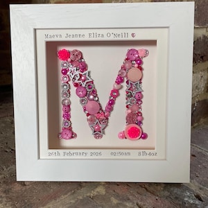 May include: A framed artwork featuring the letter "M" crafted from pink beads, buttons, and star-shaped embellishments. The frame includes the name "Maeva Jeanne Eliza O'Neill" and birth details: February 26, 2026, 02:50 am, and 8lb 4oz.