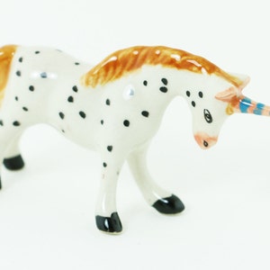 May include: A white ceramic unicorn figurine with black spots, brown mane and tail, and a blue and pink striped horn.