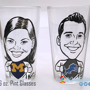 May include: Two clear 16 oz. pint glasses with hand-drawn caricatures. One glass features a woman with a University of Michigan logo, the other a man with a Detroit Lions logo. Both are in a superhero pose.