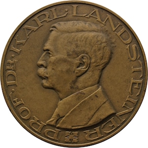 May include: A bronze coin with a profile of a man with a mustache. The text "DR. KARL LANDSTEINER" is inscribed around the edge of the coin.