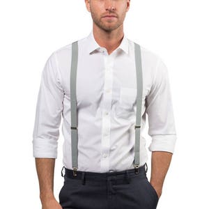 May include: A man wearing a white button-down shirt with gray suspenders and dark blue pants. He is wearing brown leather shoes.