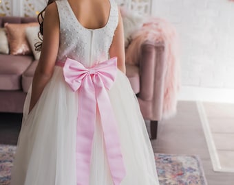 May include: A white tulle dress with a pink satin bow at the waist. The dress has a sleeveless bodice with a scoop neckline and a full skirt.
