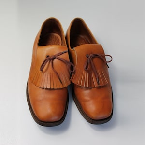 May include: A pair of brown leather tassel loafers with a fringe detail. The shoes have a rounded toe and a low heel.