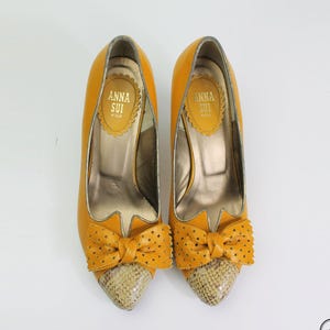 May include: A pair of yellow high heels with bow details and snakeskin-effect toes. The insoles display the brand name "ANNA SUI". The shoes are presented on a white backdrop.