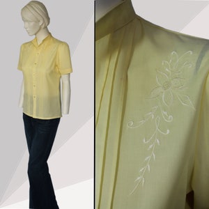 Puede incluir: Una camisa de botones de color amarillo p&aacute;lido con mangas cortas y un dise&ntilde;o floral bordado en blanco en el hombro derecho.