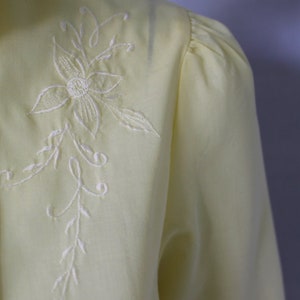 Puede incluir: Una blusa de color amarillo p&aacute;lido con un dise&ntilde;o floral bordado en blanco. El bordado presenta una flor delicada con un tallo y hojas.