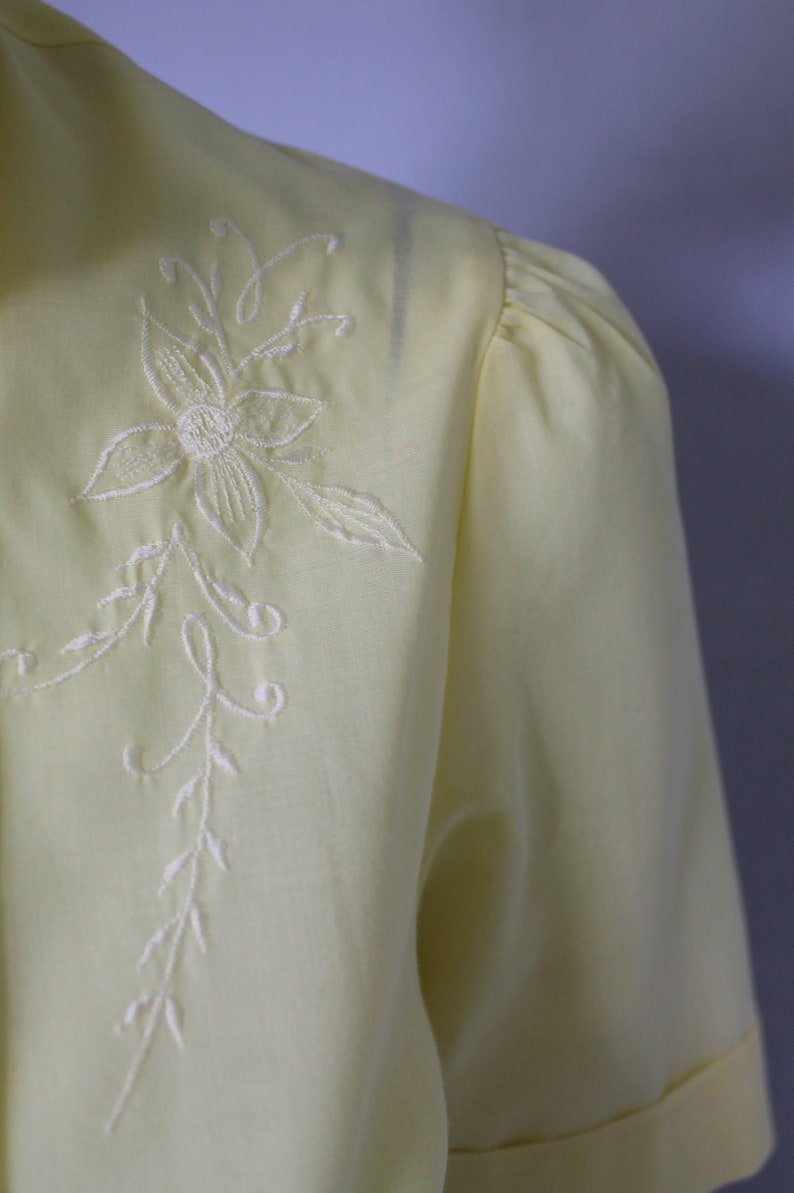 Puede incluir: Una blusa de color amarillo p&aacute;lido con un dise&ntilde;o floral bordado en blanco. El bordado presenta una flor delicada con un tallo y hojas.