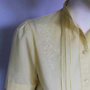 Puede incluir: Una blusa de manga corta de color amarillo p&aacute;lido con un dise&ntilde;o floral bordado en blanco en el hombro izquierdo. La blusa tiene un cierre de botones en la parte delantera y un detalle plisado en la parte delantera.
