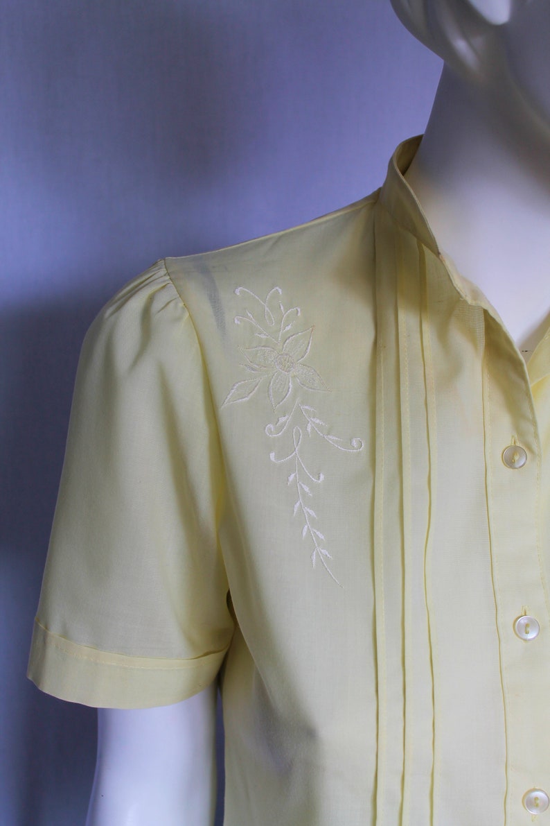Puede incluir: Una blusa de manga corta de color amarillo p&aacute;lido con un dise&ntilde;o floral bordado en blanco en el hombro izquierdo. La blusa tiene un cierre de botones en la parte delantera y un detalle plisado en la parte delantera.