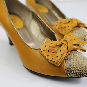 May include: Mustard yellow pumps with a bow detail. The shoes have a snakeskin pattern on the toe and a small heel. The bow features star-shaped cutouts. The interior of the shoe has a gold trim.