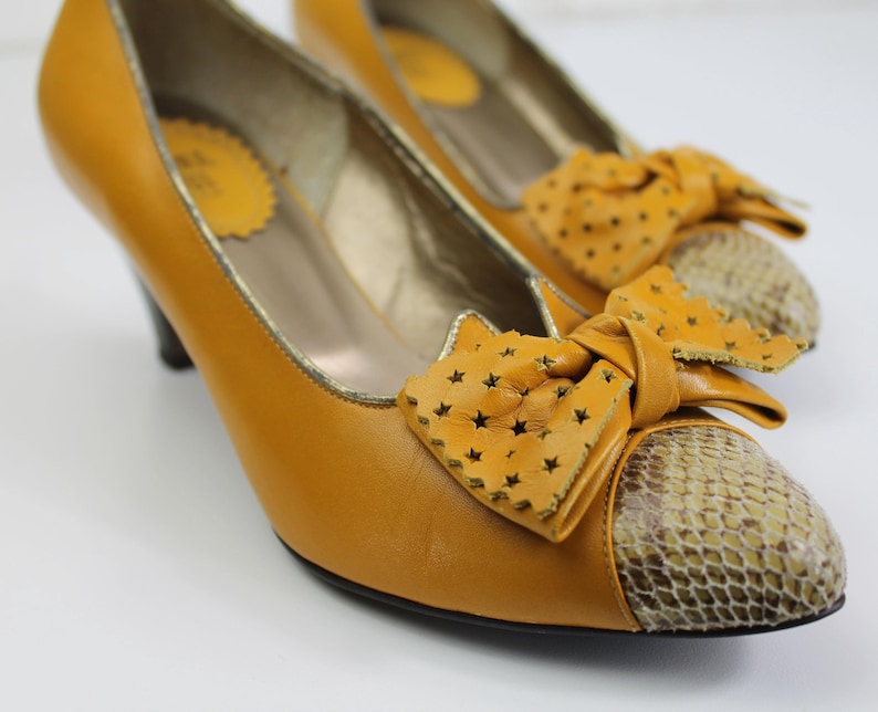 May include: Mustard yellow pumps with a bow detail. The shoes have a snakeskin pattern on the toe and a small heel. The bow features star-shaped cutouts. The interior of the shoe has a gold trim.
