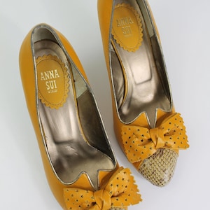 May include: A pair of mustard yellow high heels with a bow detail. The bow is adorned with star cutouts and sits atop a snakeskin-patterned toe. The interior of the shoe has a gold metallic finish. The brand name "ANNA SUI" is visible.