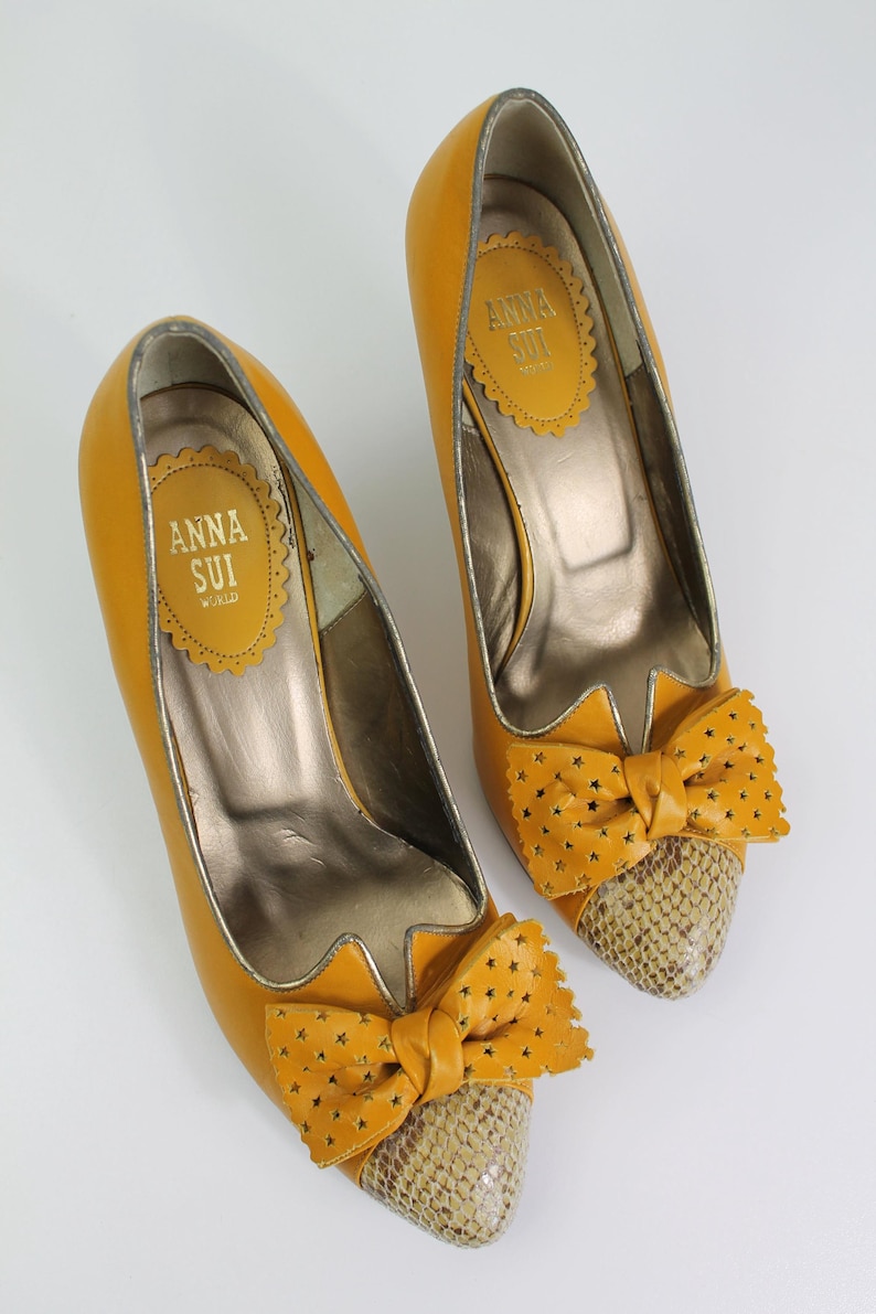 May include: A pair of mustard yellow high heels with a bow detail. The bow is adorned with star cutouts and sits atop a snakeskin-patterned toe. The interior of the shoe has a gold metallic finish. The brand name "ANNA SUI" is visible.
