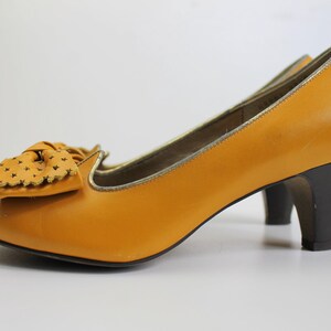 May include: Mustard yellow pumps with a bow detail and a snakeskin toe cap. The heels are black and the shoes have a gold trim. These vintage-style heels are a classic addition to any wardrobe.