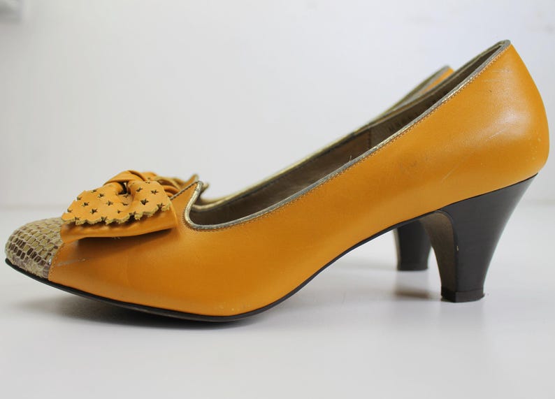 May include: Mustard yellow pumps with a bow detail and a snakeskin toe cap. The heels are black and the shoes have a gold trim. These vintage-style heels are a classic addition to any wardrobe.