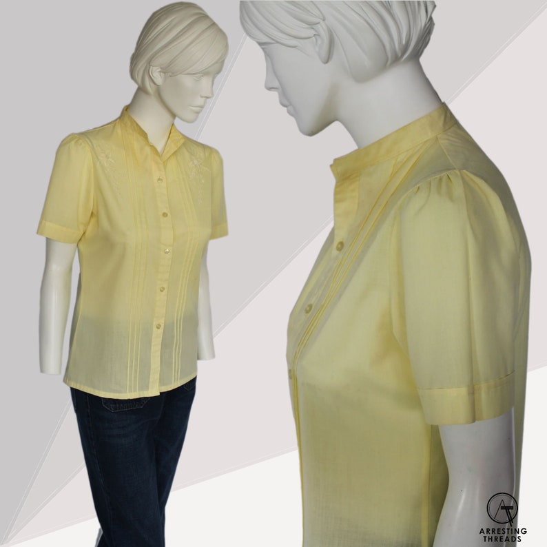 Puede incluir: Una camisa de color amarillo p&aacute;lido con botones, mangas cortas y cuello. La camisa tiene pliegues verticales en la parte delantera y un peque&ntilde;o dise&ntilde;o bordado en el cuello.
