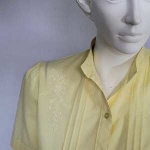 Puede incluir: Una camisa amarilla con botones y un dise&ntilde;o floral bordado en blanco en el lado izquierdo. La camisa tiene cuello y mangas cortas.
