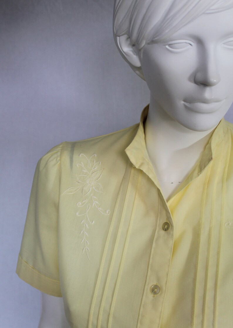 Puede incluir: Una camisa amarilla con botones y un dise&ntilde;o floral bordado en blanco en el lado izquierdo. La camisa tiene cuello y mangas cortas.