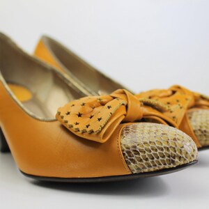 May include: A pair of mustard-colored high heels with a bow detail. The heels have a snakeskin pattern on the toe and a decorative bow with star-shaped cutouts. The shoes have a small heel.