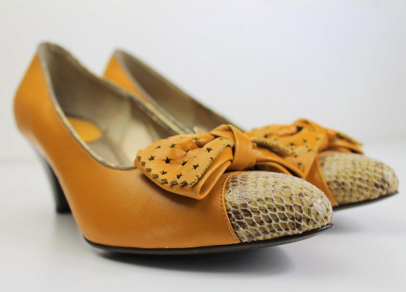 May include: A pair of mustard-colored high heels with a bow detail. The heels have a snakeskin pattern on the toe and a decorative bow with star-shaped cutouts. The shoes have a small heel.