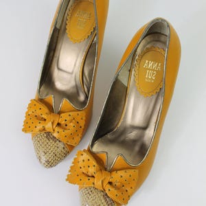 May include: A pair of mustard yellow high heels with bow accents. The heels have a snakeskin pattern on the toe and a star-shaped cutout bow. The interior of the shoe has a gold metallic finish, and the brand name "ANNA SUI" is visible.