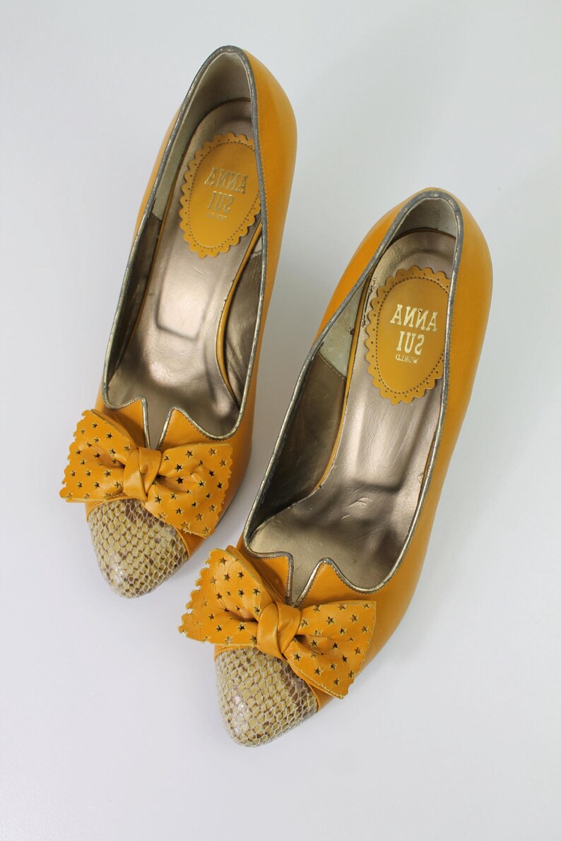 May include: A pair of mustard yellow high heels with bow accents. The heels have a snakeskin pattern on the toe and a star-shaped cutout bow. The interior of the shoe has a gold metallic finish, and the brand name "ANNA SUI" is visible.