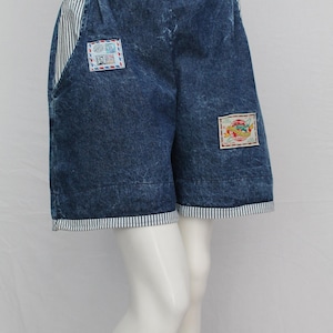 May include: Blue denim shorts with a faded wash, featuring striped accents on the pockets and hem. Decorative patches with colorful designs are sewn onto the front. The shorts have an elastic waistband.