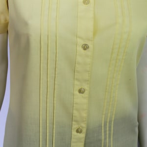 Puede incluir: Una camisa de color amarillo p&aacute;lido con botones, cuello y mangas cortas. La camisa tiene pliegues verticales y una fila de botones en la parte delantera.