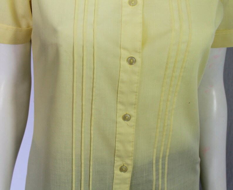 Puede incluir: Una camisa de color amarillo p&aacute;lido con botones, cuello y mangas cortas. La camisa tiene pliegues verticales y una fila de botones en la parte delantera.