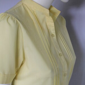 Puede incluir: Una blusa de manga corta de color amarillo p&aacute;lido con cuello mandar&iacute;n y una fila de botones en la parte delantera. La blusa tiene un frente plisado y est&aacute; hecha de una tela ligera.